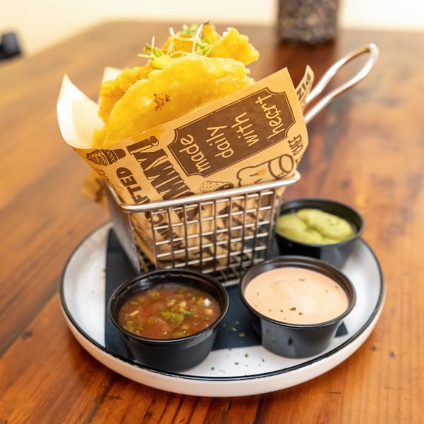Golden fried Tostao's tapas in a metal basket with three dipping sauces on a white plate.