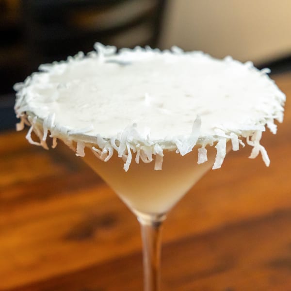 A creamy Tostao's cocktail in a martini glass, rimmed with shredded coconut, on a wooden table.